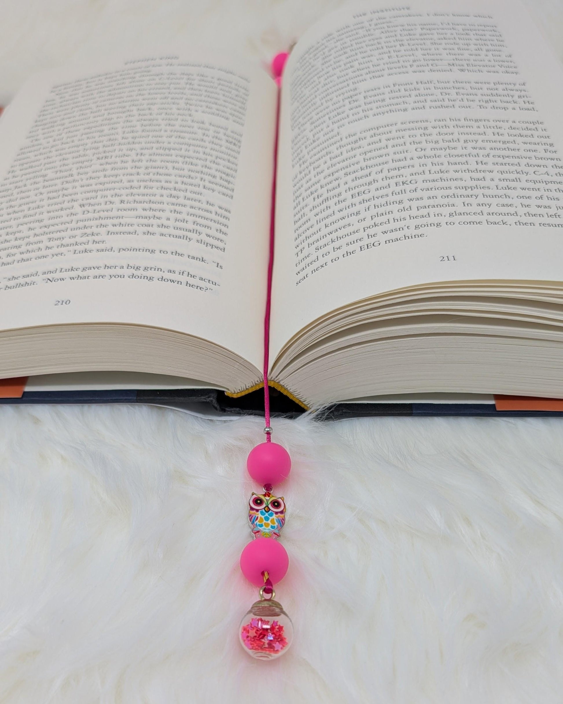 May include: A pink bookmark with a beaded charm. The charm includes a pink bead, a colorful owl bead, another pink bead, and a clear glass jar with pink glitter inside. The bookmark is placed in an open book with text on the pages.