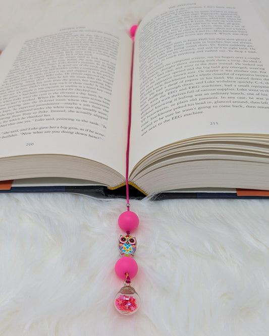 May include: A pink bookmark with a beaded charm. The charm includes a pink bead, a colorful owl bead, another pink bead, and a clear glass jar with pink glitter inside. The bookmark is placed in an open book with text on the pages.